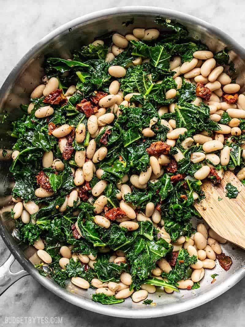 A skillet full of Sun Dried Tomato, Kale, and White Beans, with a wooden spatula.