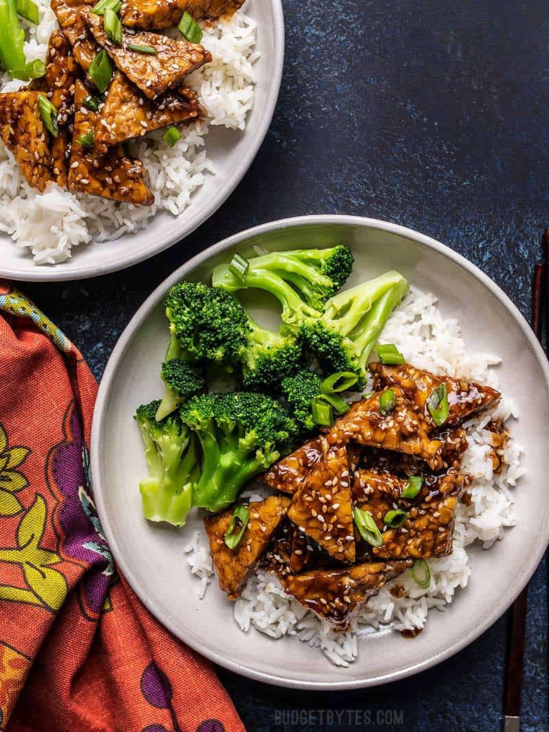 Two plates with Sesame Tempeh, rice, and broccoli, next to a colorful napkin