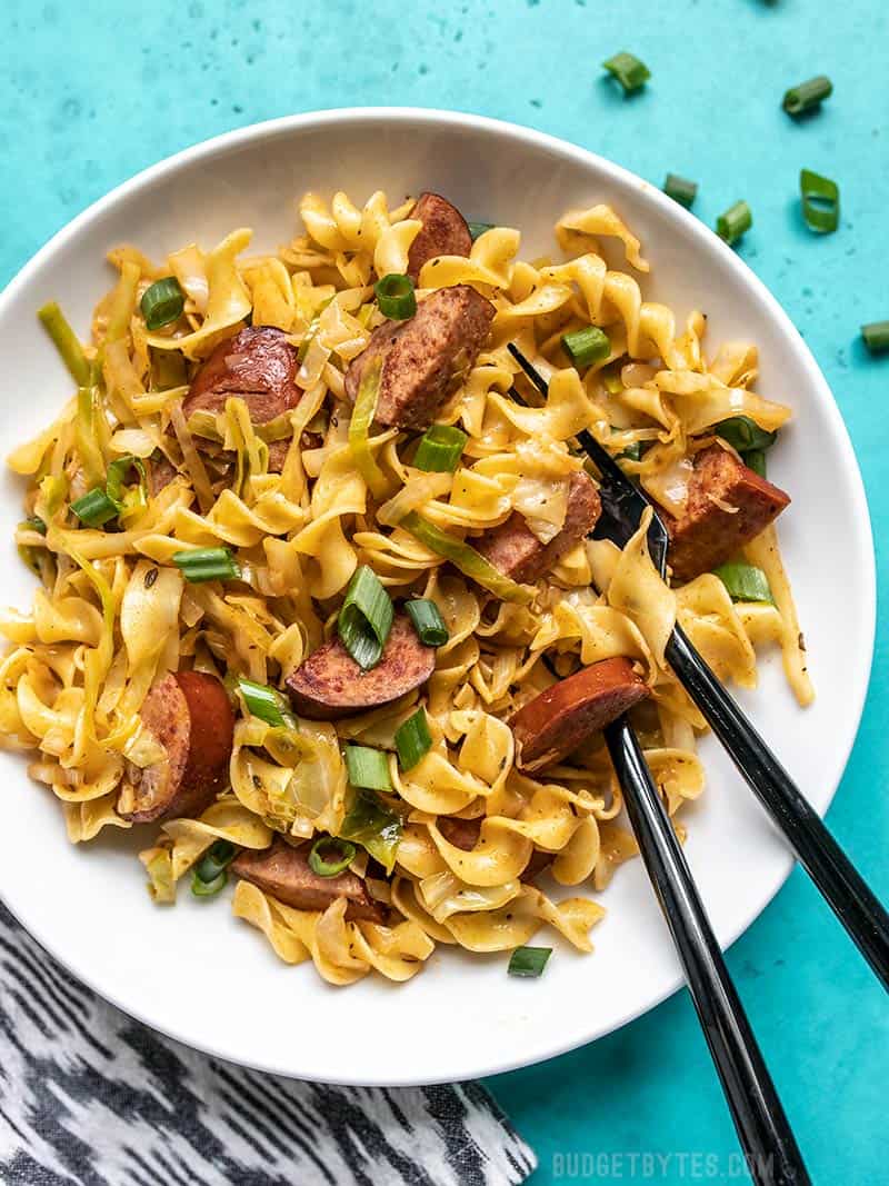 A bowl of Cajun Cabbage and Noodles with a black spoon and fork stuck in the side of the bowl. Green onions sprinkled on top.