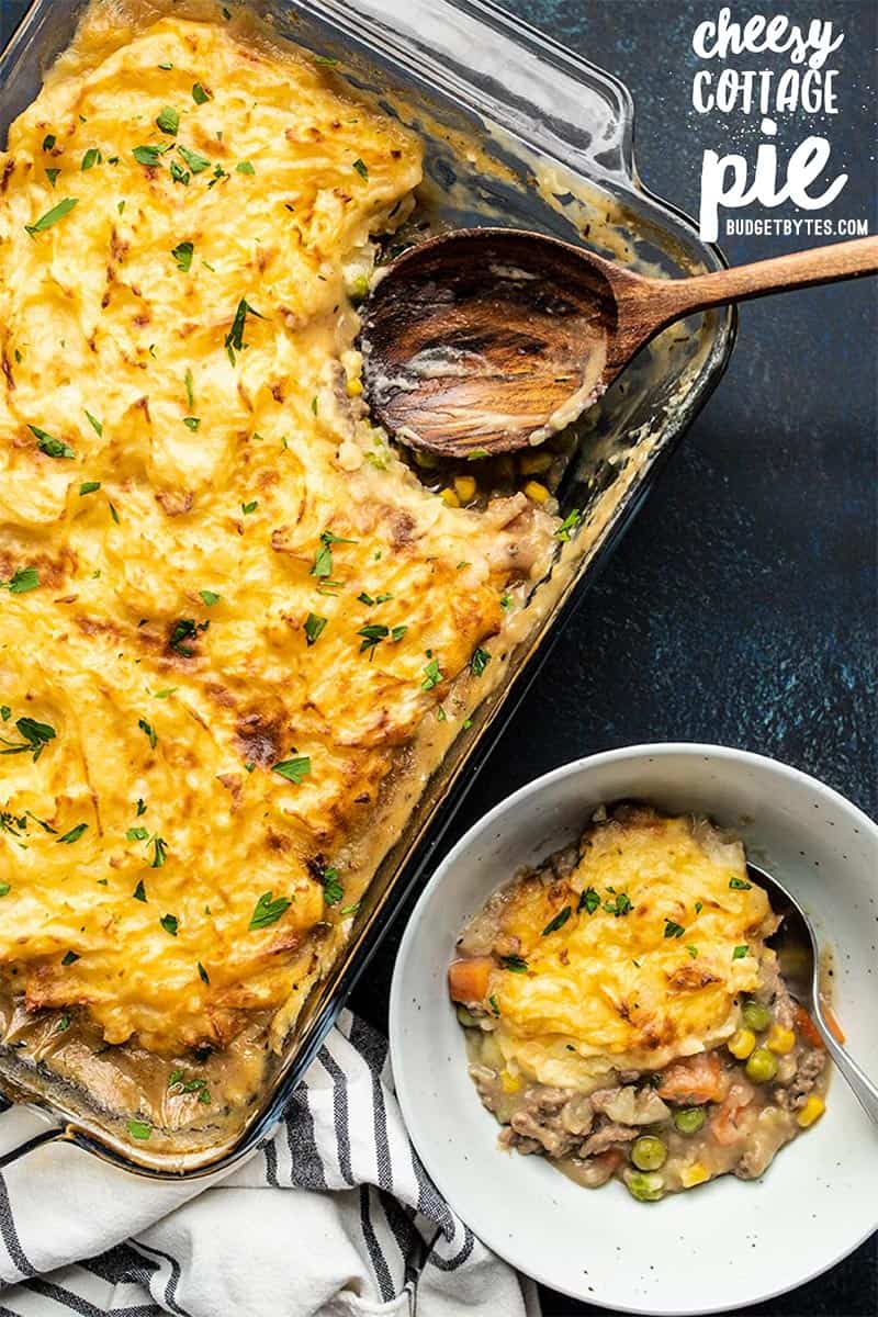 A casserole dish full of Cheesy Cottage Pie with one portion scooped out into a bowl on the side.