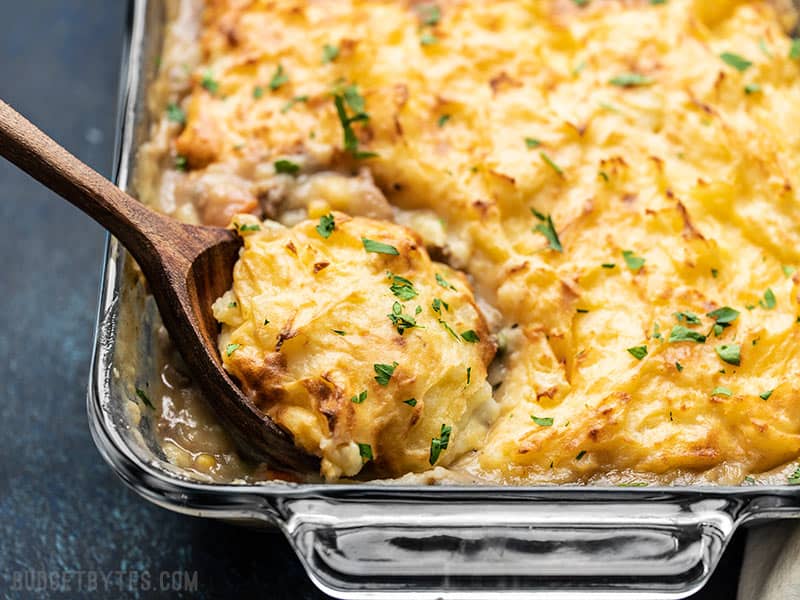 Front View of Cheesy Cottage Pie being scooped out of the glass casserole dish with a large wooden spoon