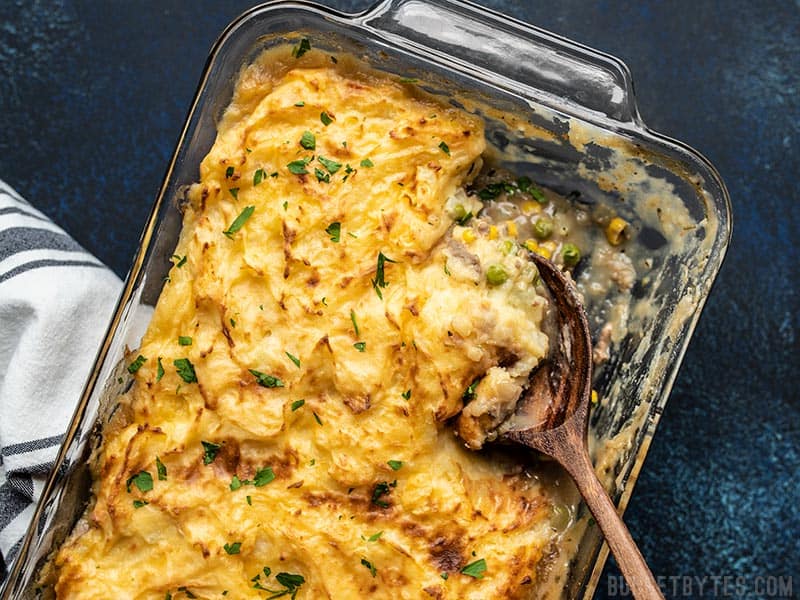 Cheesy Cottage Pie being scooped out of a glass casserole dish with a big wooden spoon. 