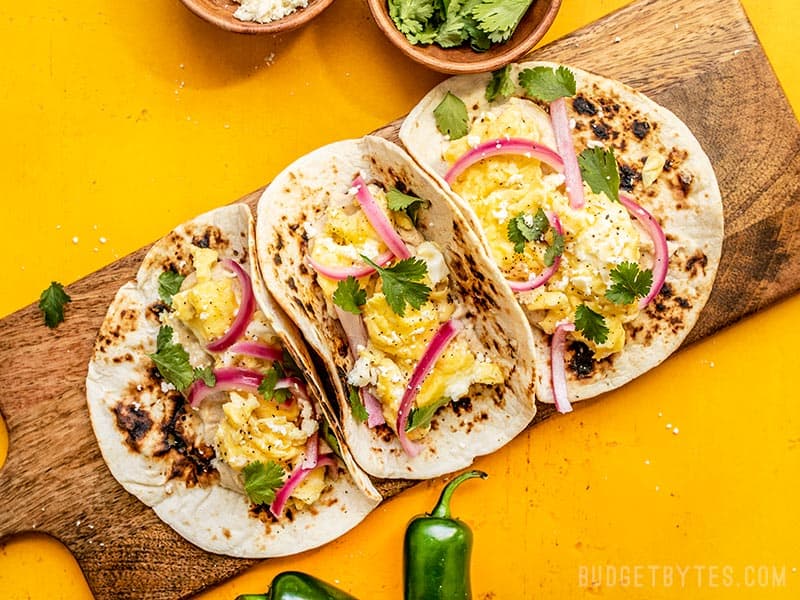 Three Hummus Breakfast Tacos on a wooden cutting board with cilantro, cheese, and jalapeños on the sides