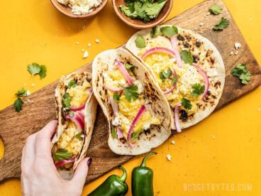 Three Hummus Breakfast Tacos on a wooden cutting board, a hand picking one up