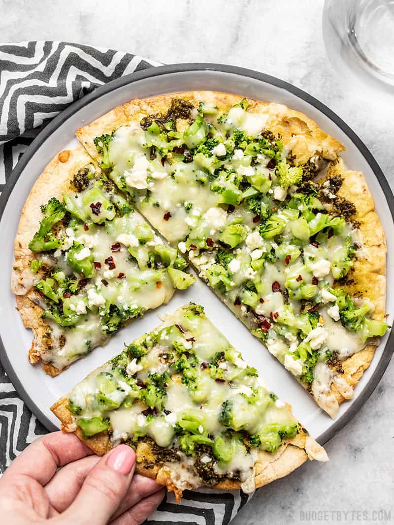 A Quick Fix Broccoli Pesto Pizza on a plate with a striped napkin and a hand pulling one slice away. 