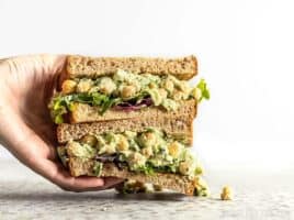 A hand grabbing a stack of two halves of a Scallion Herb Chickpea Salad sandwich on wheat bread.