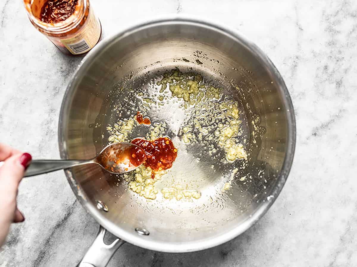 Chili sauce being added to a sauce pot with ginger and garlic. 