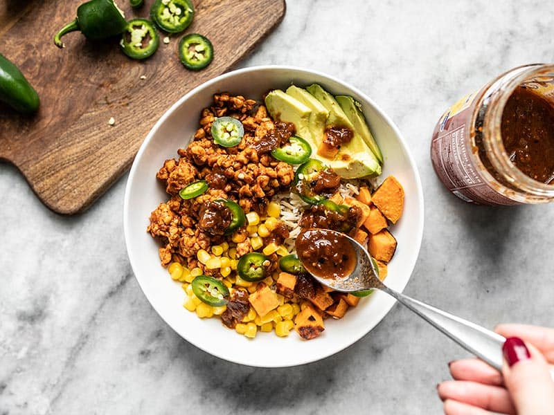 Salsa being spooned onto a tempeh burrito bowl with sliced jalapeños on the side