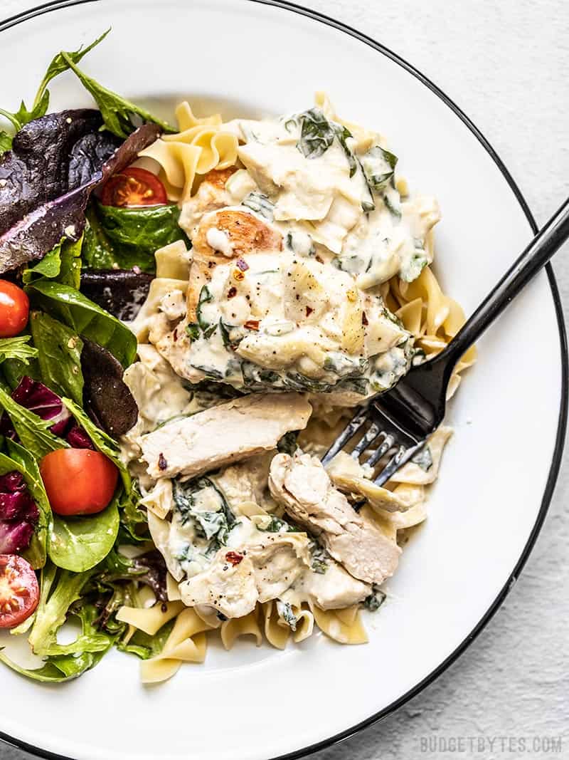 Creamy Spinach Artichoke Chicken on a plate with a bed of pasta and a salad on the side