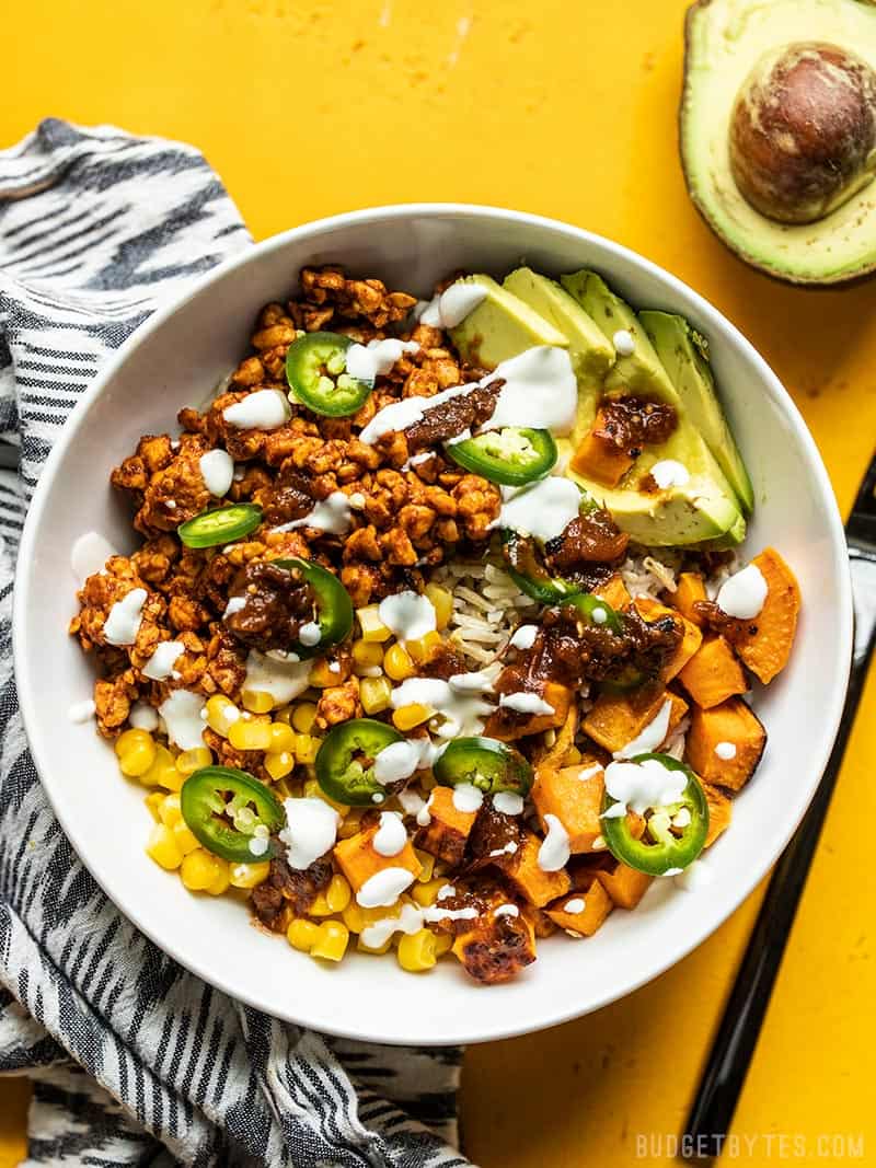 Close up overhead view of a Tempeh Burrito Bowl on a yellow background and an avocado on the side.