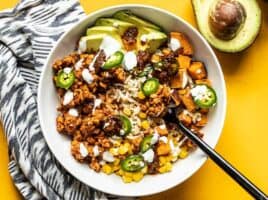 Overhead view of a tempeh burrito bowl with a black fork digging in, an avocado on the side.