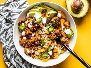 Overhead view of a tempeh burrito bowl with a black fork digging in, an avocado on the side.