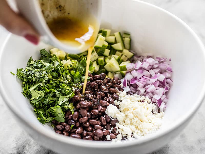 Dressing being poured onto cucumber and black bean salad ingredients