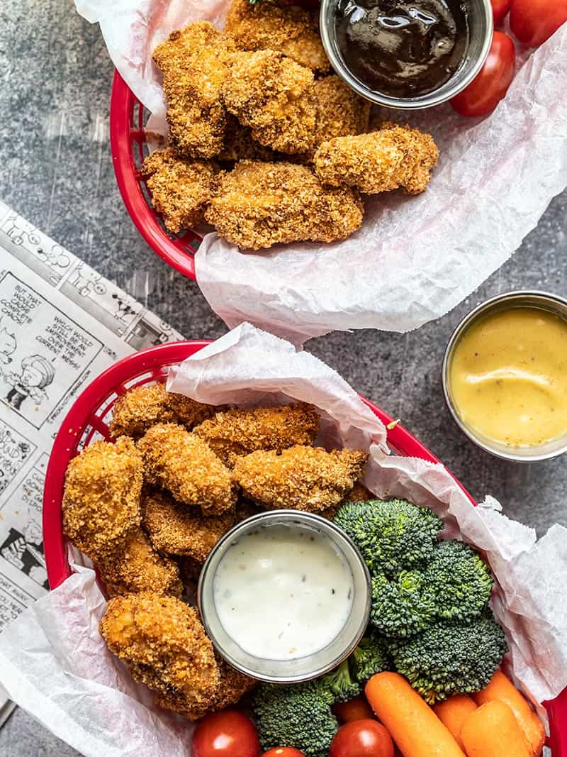 Two baskets with Homemade Baked Chicken Nuggets, vegetables, and dip sitting next to a newspaper.