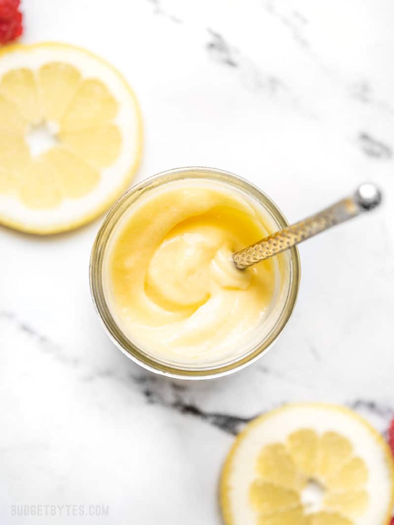 Overhead view of a jar of homemade lemon curd with a butter knife inside and slices of lemon on the sides.