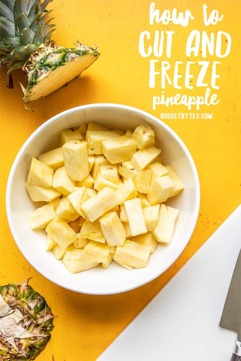 A bowl of cut pineapple next to a cutting board and the pineapple top.