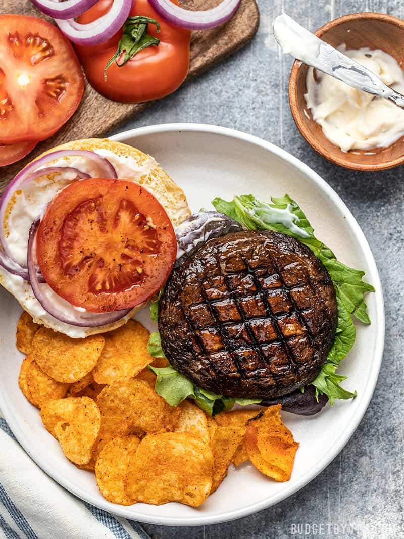 An open Marinated Portobello Mushroom Burger on a plate with chips, mayo, sliced tomato, and red onion rings on the side.