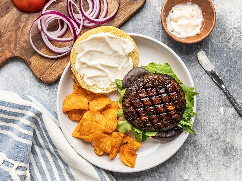 An open portobello mushroom burger on a plate with chips, a small dish of mayo and onion rings on the side.