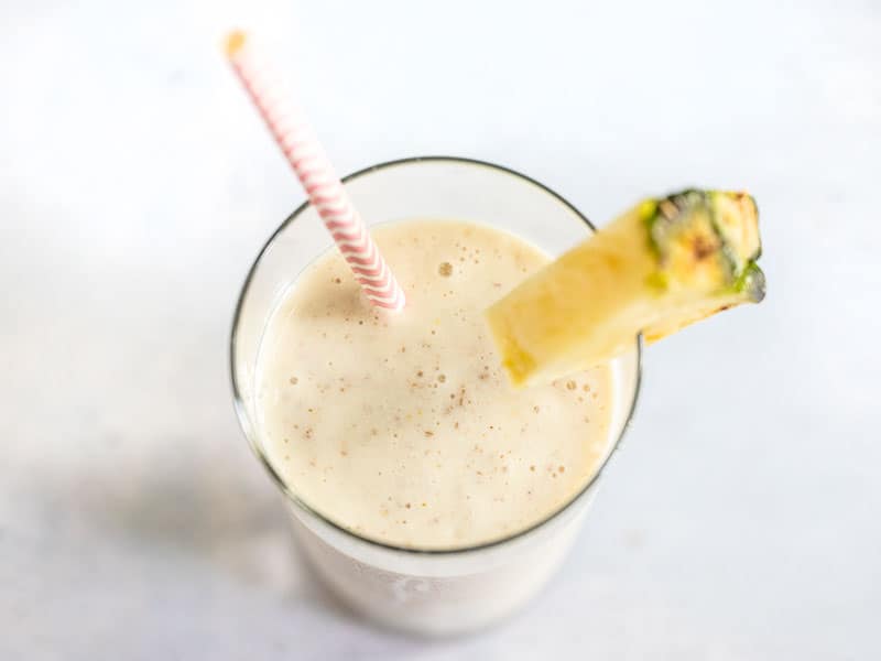 Close up of Pineapple Protein Smoothie in the glass, with a paper straw and fresh pineapple wedge on the rim.