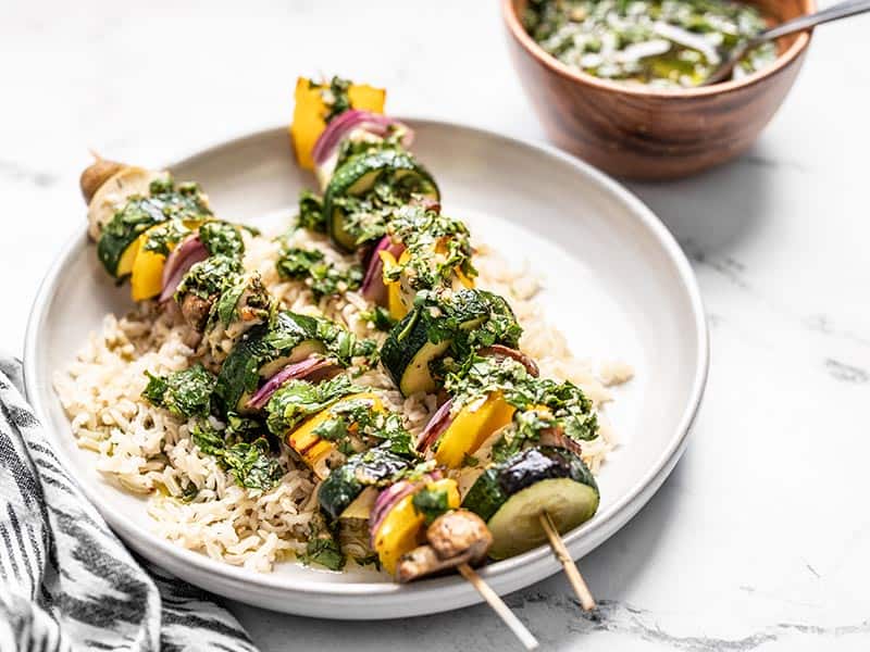 Front view of a plate with rice and two chimichurri chicken kebabs, with a wooden bowl of chimichurri in the back.