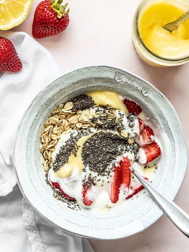 A half-stirred lemon berry yogurt breakfast bowl with a jar of lemon curd and some strawberries on the side.