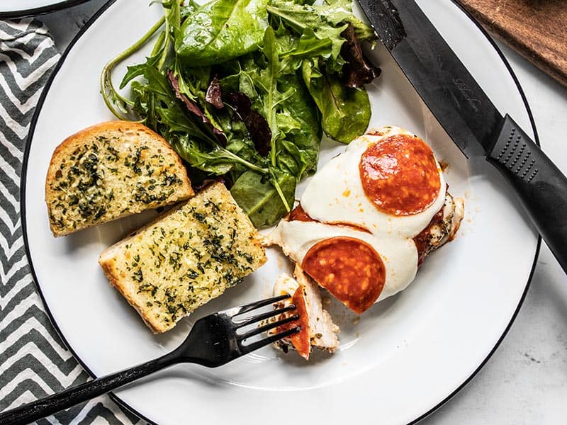 A fork taking a piece of Baked Pizza Chicken from the plate with chicken, garlic bread, and salad.