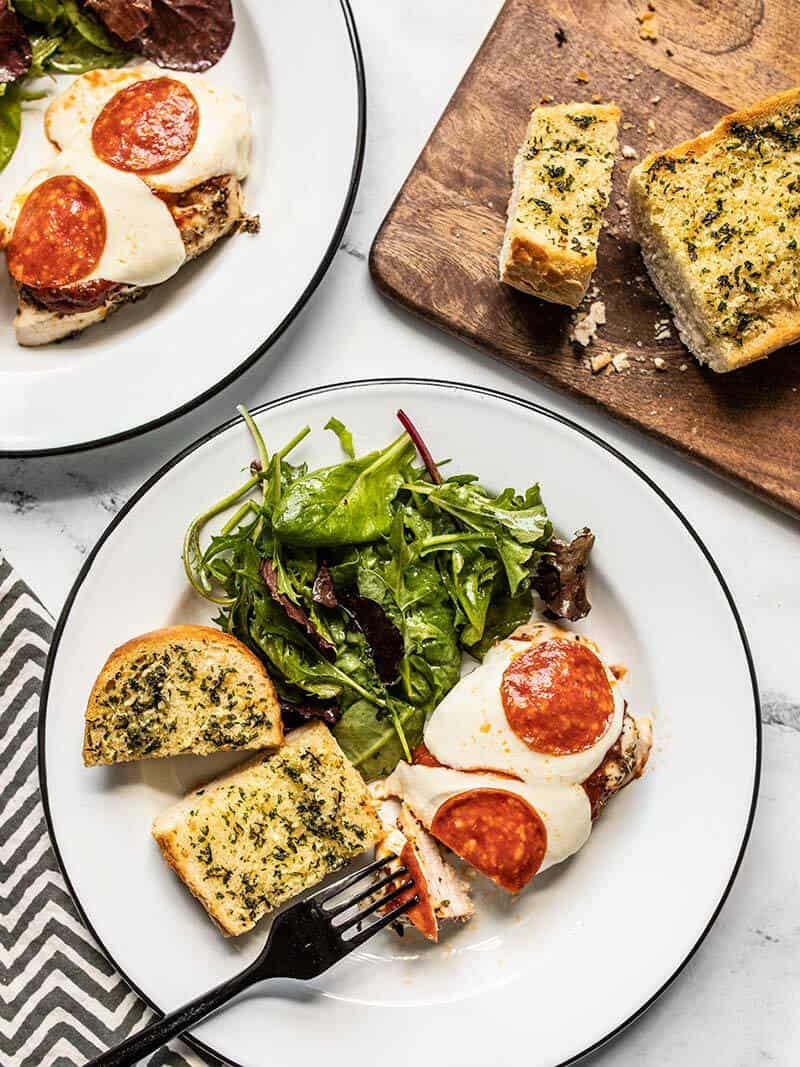 Two plates with Baked Chicken Pizza, salad, and garlic bread next to a cutting board with more garlic bread.