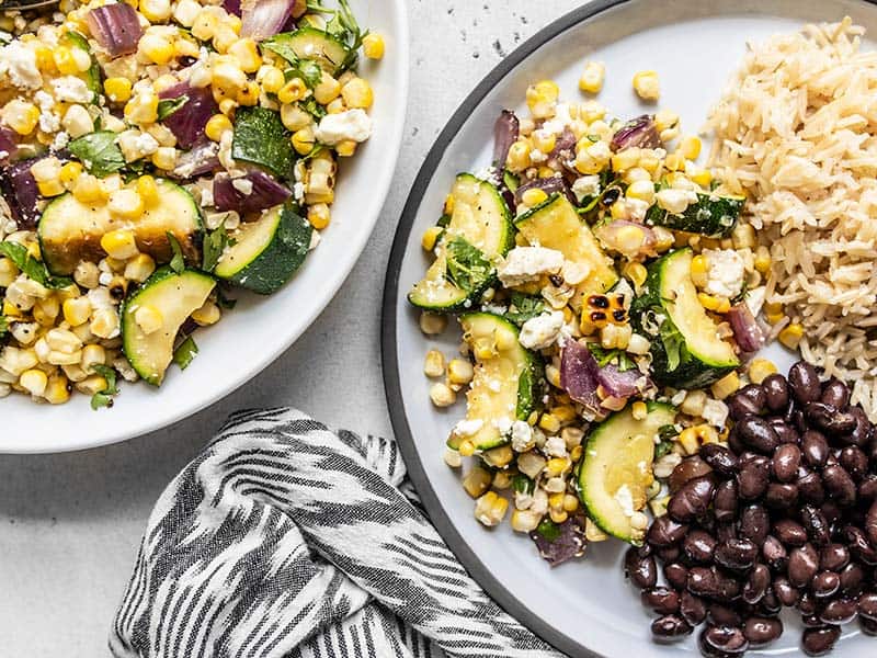 A plate filled with Charred Corn and Zucchini Salad, black beans, and rice, next to a bowl of the corn and zucchini salad.