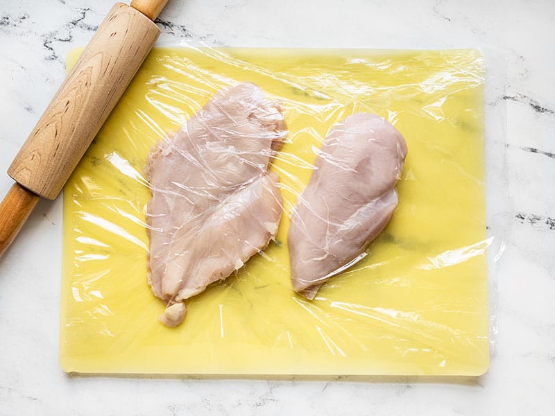 Chicken breasts on a cutting board covered with plastic with a rolling pin on the side.