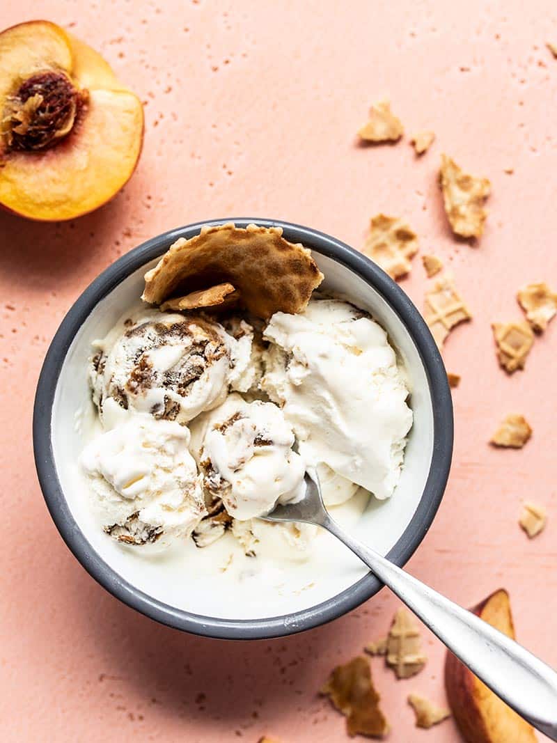 One bowl of No Churn Balsamic Peach Ice Cream with a fresh peach on the side and pieces of waffle cone in the bowl and on the counter.