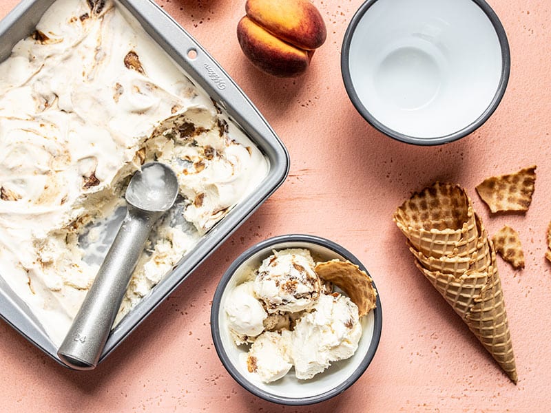 Baking pan full of No Churn Balsamic Peach Ice Cream scooped out and served in one bowl, with one empty bowl, and some waffle cones.