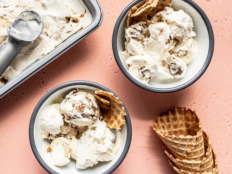 Two bowls of No Churn Balsamic Peach Ice Cream with the pan of ice cream and a stack of waffle cones on the side.