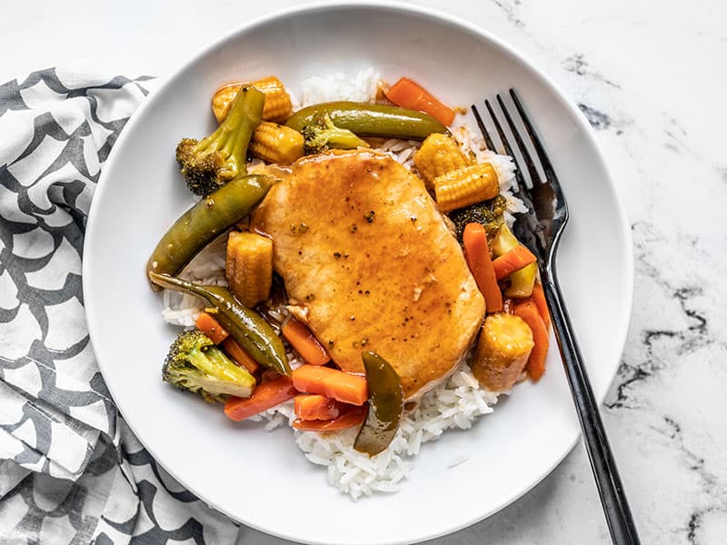 A plate with rice and sweet and sour pork chops with vegetables, a black fork on the side of the plate.