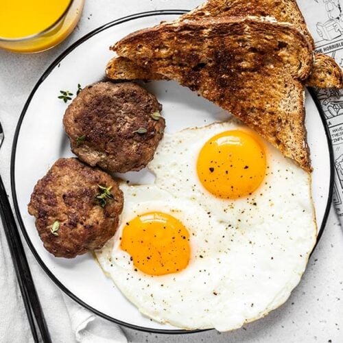 A breakfast plate with eggs, toast, and maple sage breakfast sausage next to a newspaper and glass of orange juice.