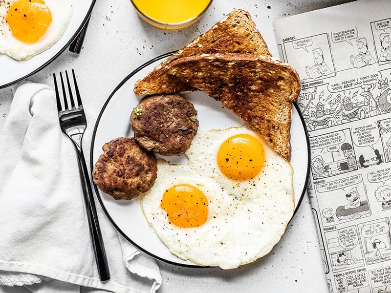 A breakfast plate with eggs, toast, and maple sage breakfast sausage, next to a newspaper and glass of orange juice.