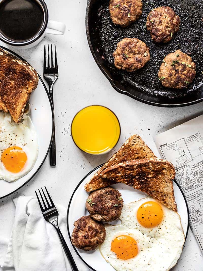 Tablescape with two plates full of eggs, toast, and maple sage breakfast sausage, a cast iron skillet with sausage, and drinks