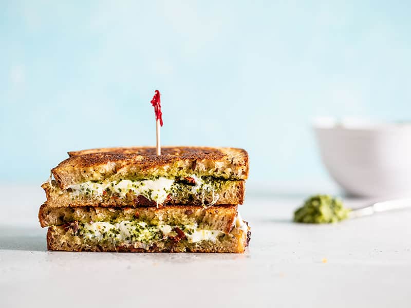 Two stacked pieces of pesto grilled cheese next to a bowl and small knife with pesto on it.