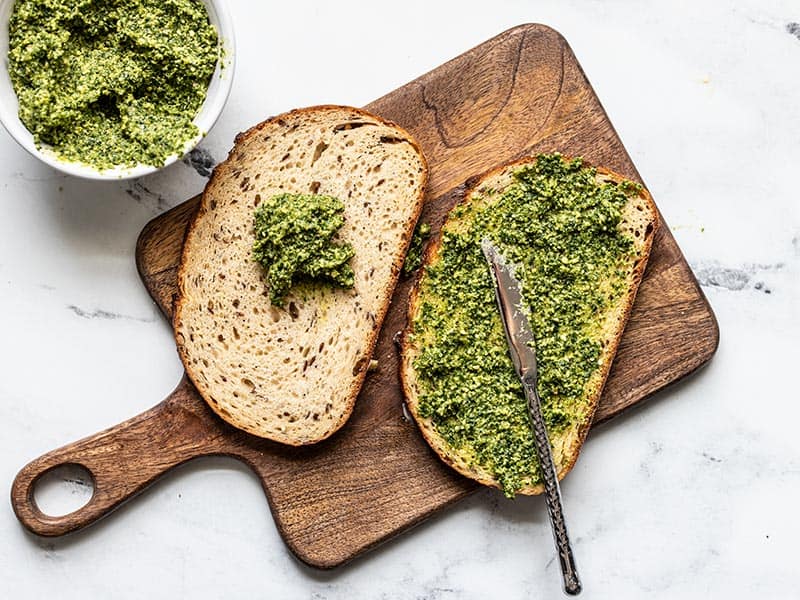 Pesto being spread onto two slices of bread, a bowl of pesto on the side.