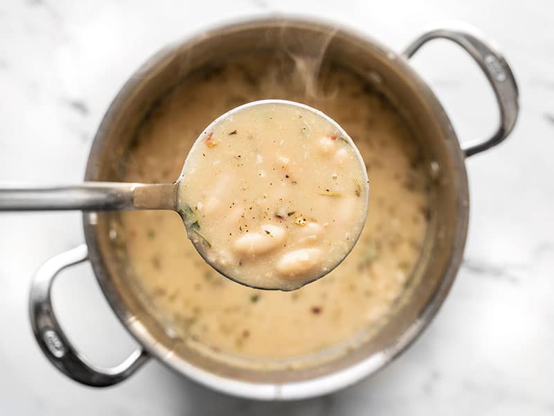 Close up of a ladle full of rosemary garlic white bean soup hovering over the soup pot, steam coming off the soup.