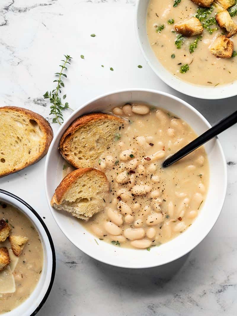 Three bowls of Rosemary Garlic White Bean Soup with different toppings like toasted bread, croutons, and pesto.