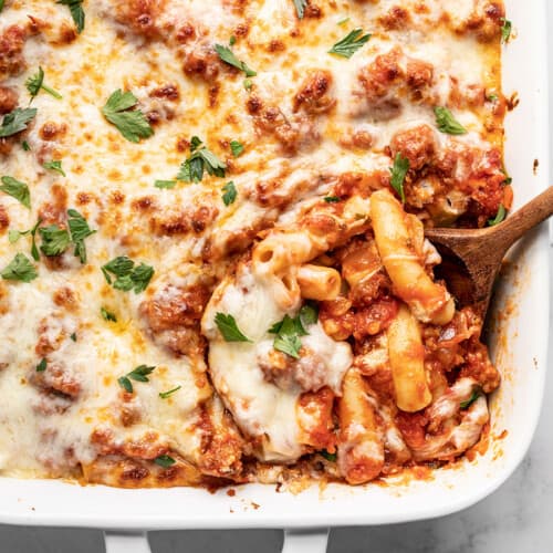 Overhead view of baked ziti being scooped out of the casserole dish with a wooden spoon.