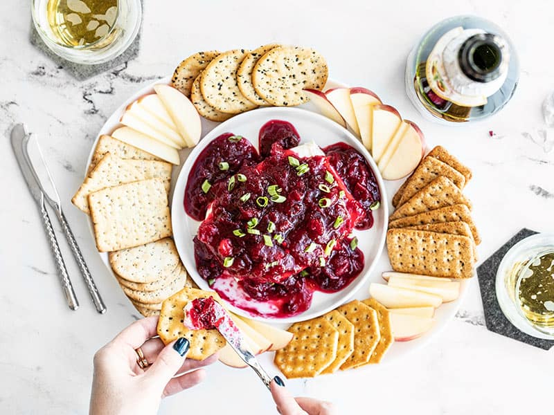 Two hands smearing cranberry cream cheese dip on a cracker over the platter