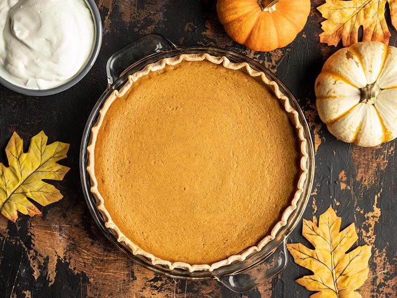 Overhead view of a baked Maple Brown Butter Pumpkin Pie, uncut with whipped cream, mini pumpkins, and leaves on the side.