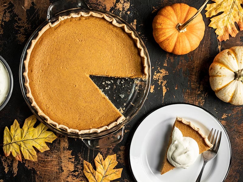 Overhead view of a Maple Brown Butter Pumpkin Pie with a slice cut out, on a plate on the side with whipped cream.