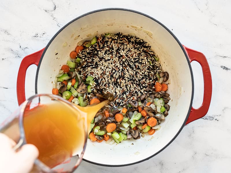 Vegetable broth being poured into the pot with vegetables and wild rice