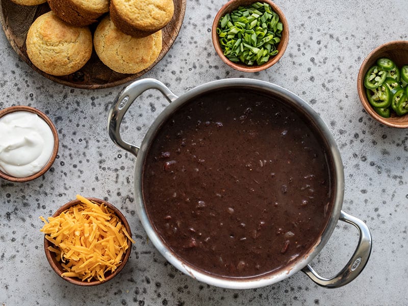 Pot of Smoky Black Bean Soup surrounded by toppings like cheddar, sour cream, green onion, and jalapeño