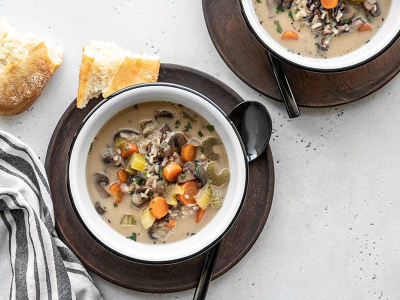 Two bowls of creamy vegetable wild rice soup on wooden plates, each with a piece of bread on the side