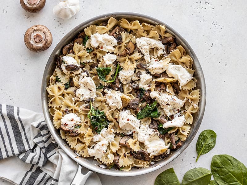 Finished Mushroom and Spinach Pasta with Ricotta in the skillet next to spinach leaves and whole mushrooms