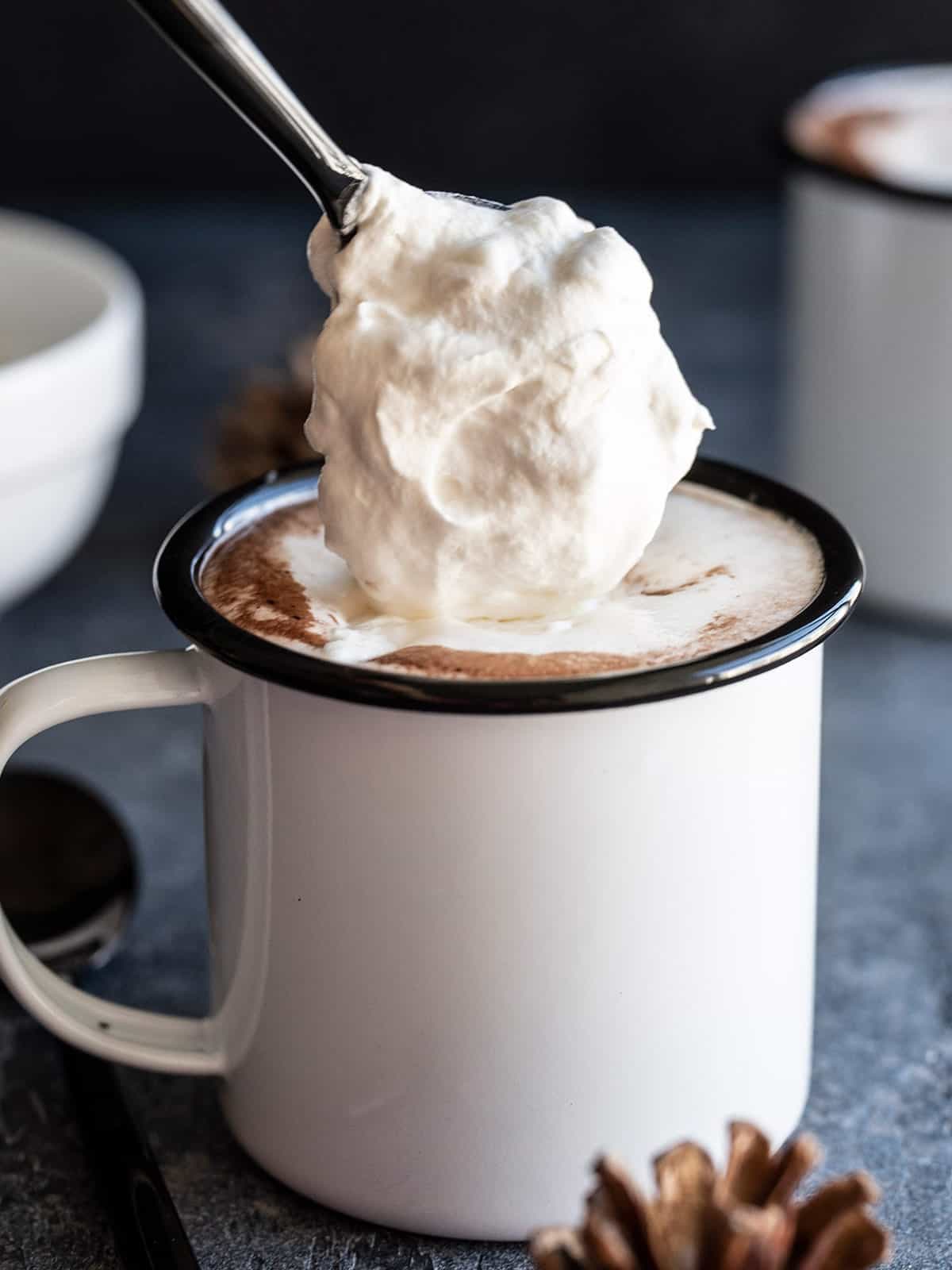 Side view of whipped cream being spooned onto a mug of hot chocolate.