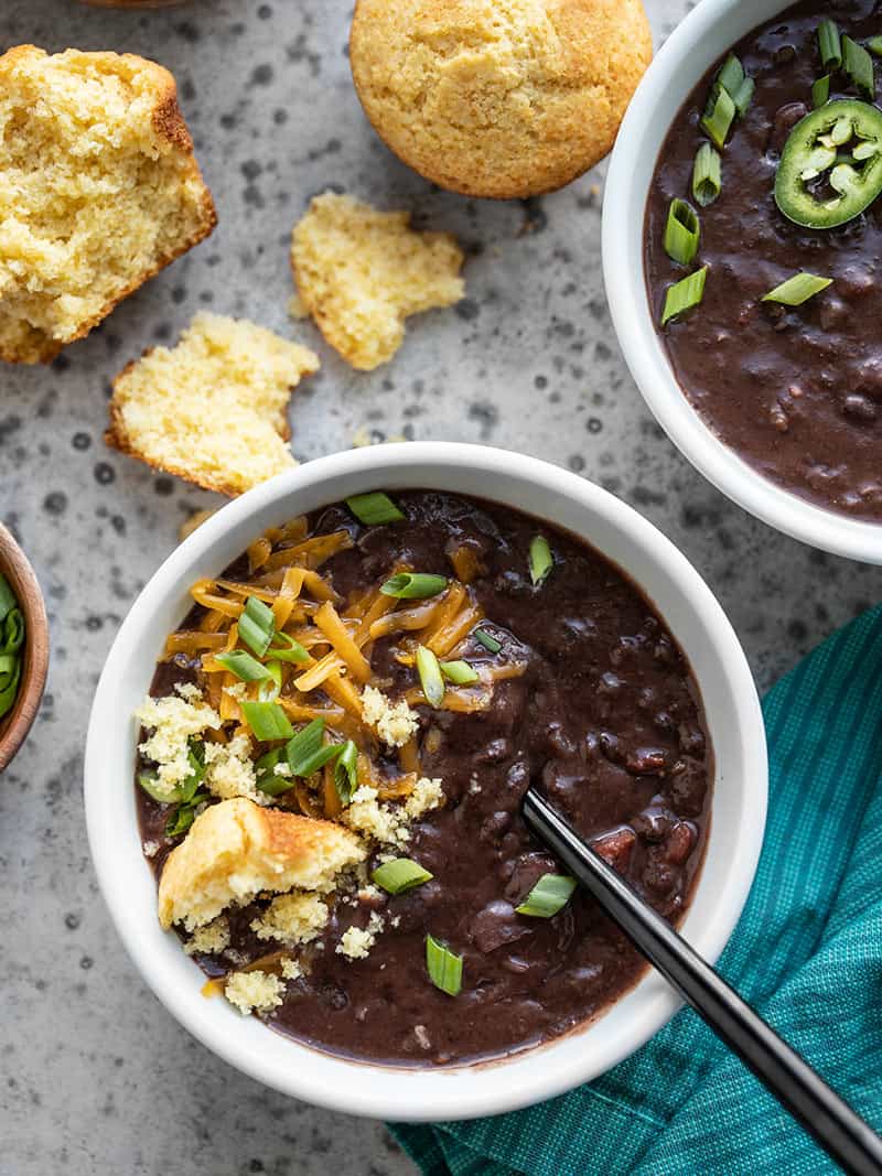 Two bowls of Smoky Black Bean Soup with different toppings, like cornbread, jalapeño, cheddar, and green onion